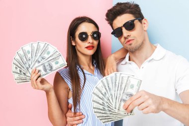 Heyecanlı genç çift ayakta para banknot gösterilen, güneş gözlüğü takıyor iki renkli arka plan, izole