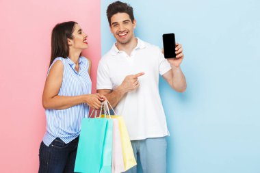 Mutlu genç çift ayakta alışveriş çantaları taşırken, boş ekran cep telefonu gösterilen iki renkli arka plan, izole