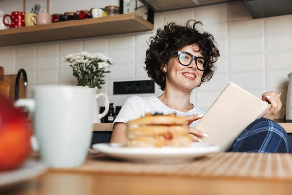 微笑着的快乐女孩 坐在家里的厨房里看书 吃美味的早餐 — 图库照片