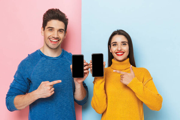 Beautiful young couple standing isolated over two colored background, showing blank screen mobile phones