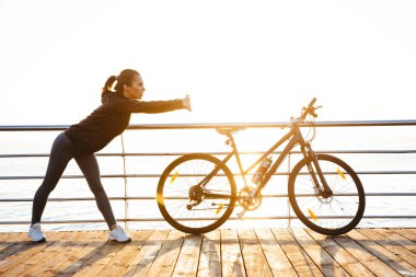 Okyanus üzerinde gündoğumu sırasında açık havada bisiklet yakın dururken germe yapıyor atletik kadının fotoğrafını egzersizleri