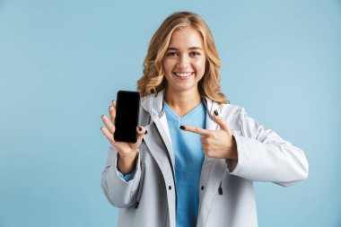 Boş ekran cep telefonu parmak işaret üzerinde mavi arka plan, izole ayakta yağmurluk giyen neşeli genç kız