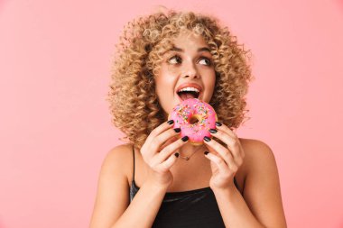 Fotoğraf eğlenceli kıvırcık kadın 20'li yaşlarda giyen elbise pembe arka plan üzerinde izole ayakta iken donut yemek