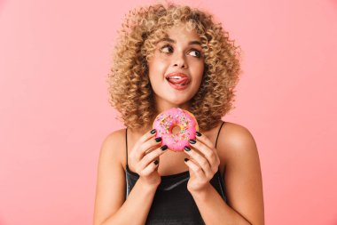 Fotoğraf sevimli kıvırcık kadın 20'li yaşlarda giyen elbise pembe arka plan üzerinde izole ayakta iken donut yemek