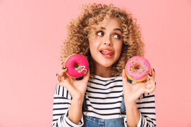 Çok kıvırcık kadın 20'li yaşlarda giyiyor rahat kıyafetler donuts pembe arka plan üzerinde izole ayakta süre tutarak fotoğrafı