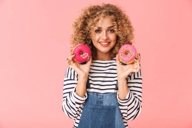 Sevimli kıvırcık kadın 20'li yaşlarda giyiyor rahat kıyafetler donuts pembe arka plan üzerinde izole ayakta süre tutarak fotoğrafı