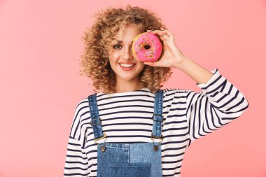 Pembe arka plan üzerinde izole ayakta iken yeme çörek görüntü olumlu kıvırcık kadın rahat giyen 20'li elbise