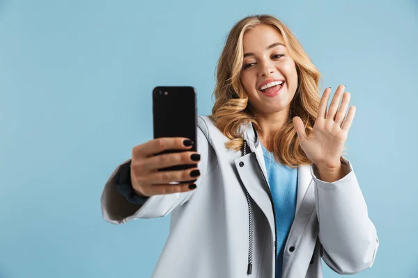 Cheerful young girl wearing raincoat standing isolated over blue background, taking a selfie