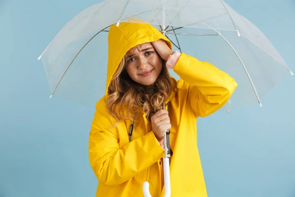 青い背景に分離した透明の傘の下で女性 代着て黄色いレインコートを着て立っている笑顔のイメージ — ストック写真
