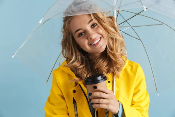 Photo of content woman 20s wearing yellow raincoat standing under transparent umbrella with takeaway coffee isolated over blue background