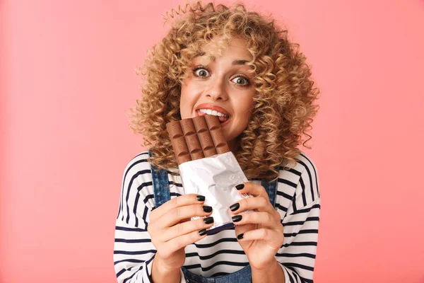 Photo of charming curly woman 20s eating chocolate bar while standing isolated over pink background