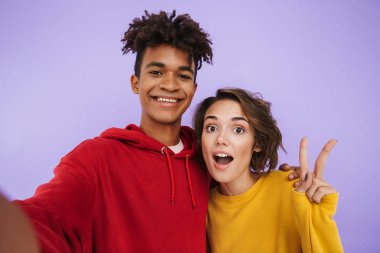 Neşeli multhiethnic genç çift ayakta birlikte bir selfie alarak, menekşe arka plan üzerinde izole