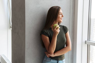 Evde duvara ayakta, yeşil elma holding neşeli genç kadın