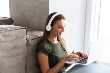 Dizüstü bilgisayarda çalışan neşeli genç kadın evde, kulaklık ile müzik dinlemek