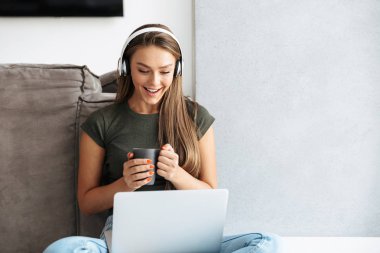 Dizüstü bilgisayar, çay içme çalışan neşeli genç kadın evde, kulaklık ile müzik dinlemek
