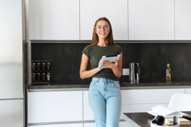 Güzel genç kadın mutfakta ayakta, tablet bilgisayar kullanarak