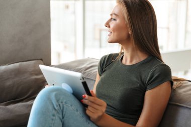 Gülümseyen genç kadın evde, bir kanepede oturan tablet bilgisayar kullanarak