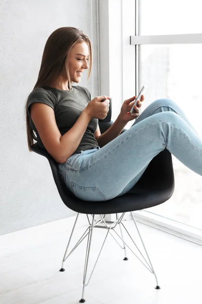 Happy young woman sitting in a chair, drinking tea, using mobile phone