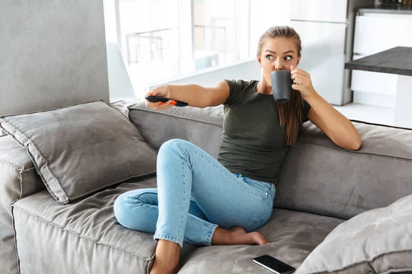 Cheerful young woman sitting on a couch at home, changing channels, drinking tea from a cup