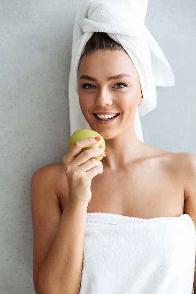 美丽的年轻女子包裹在浴巾站在家里的墙上 拿着绿色的苹果 — 图库照片