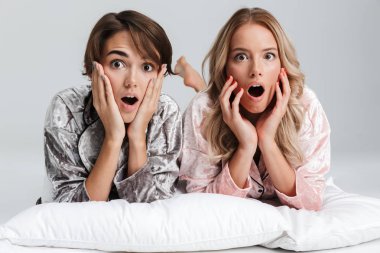 Two pretty surprised girls wearing pajamas isolated over gray background, laying on bed with pillows