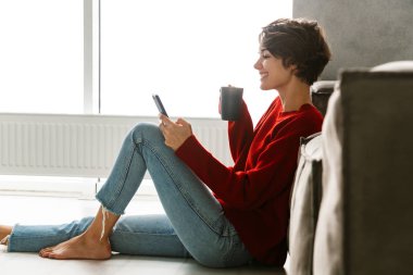 Lovely young woman wearing sweater sitting on a floor at home, drinking tea, leaning on a couch, using mobile phone