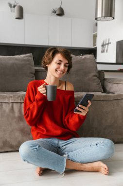 Gülümseyen kahve içme, cep telefonuyla evde, kazak giyen genç kadın