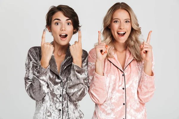 Two cheerful girls wearing pajamas standing isolated over gray background, pointing at copy space