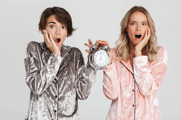 Two pretty shocked girls wearing pajamas isolated over gray background, showing alarm clock