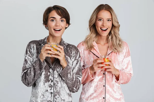 Two pretty girls wearing pajamas isolated over gray background, drinking orange juice