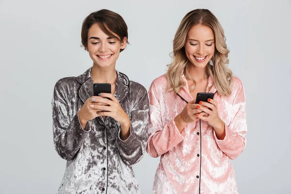 Две Симпатичные Девушки Пижаме Изолированы Сером Фоне Держат Руках Мобильные — стоковое фото