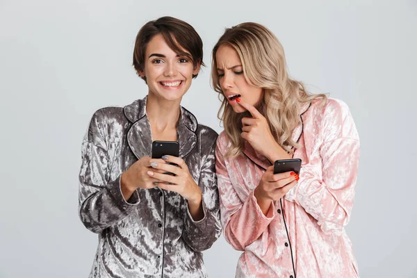 Cep telefonları tutan gri arka plan üzerinde izole pijama giymiş iki güzel kızlar