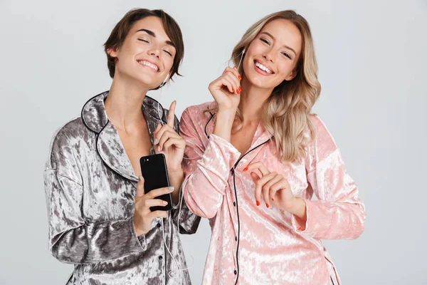 Kulaklık ve cep telefonu ile müzik dinlemek gri arka plan üzerinde izole pijama giymiş iki güzel kızlar