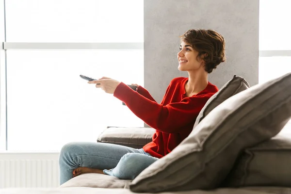 微笑的年轻女子穿着毛衣在家里的沙发上放松 喝咖啡 拿着电视遥控器 — 图库照片