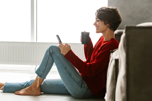 自宅の床の上に座って お茶を飲み ソファにもたれて 携帯電話を使用してのセーターを着て素敵な若い女性 — ストック写真