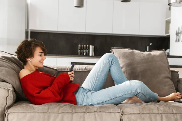 陽気な若い女性自宅でリラックス 無線イヤホンで音楽を聴く 携帯電話を保持しています — ストック写真