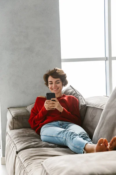 陽気な若い女性自宅でリラックス 無線イヤホンで音楽を聴く 携帯電話を保持しています — ストック写真