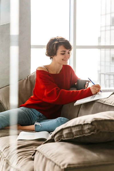 Cheerful girl studying at home while laying on a couch and drinking tea