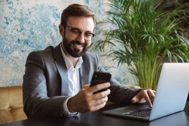 Yakışıklı genç işadamı kafede kapalı, cep telefonu tutan oturan bir dizüstü bilgisayar üzerinde çalışan takım elbise giymiş