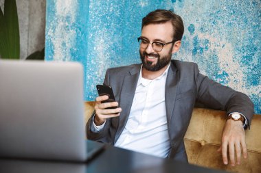 Yakışıklı genç işadamı kafede kapalı, cep telefonu tutan oturan bir dizüstü bilgisayar üzerinde çalışan takım elbise giymiş