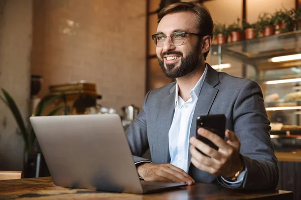 Yakışıklı genç işadamı kafede kapalı, cep telefonu tutan oturan bir dizüstü bilgisayar üzerinde çalışan takım elbise giymiş