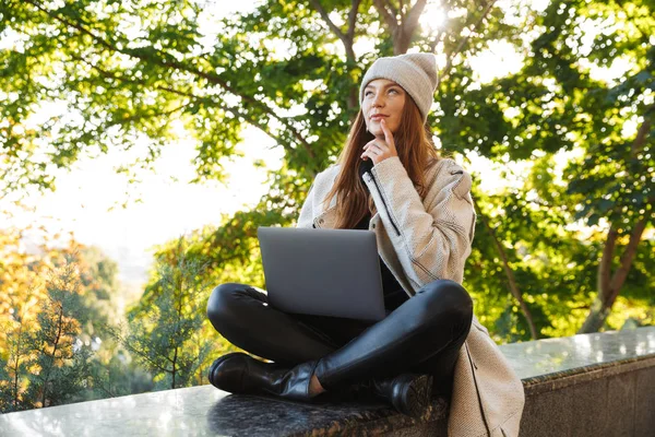 ラップトップ コンピューターを使用して 若い女性を物思いにふける秋コートと屋外で 座っている帽子に身を包んだ — ストック写真