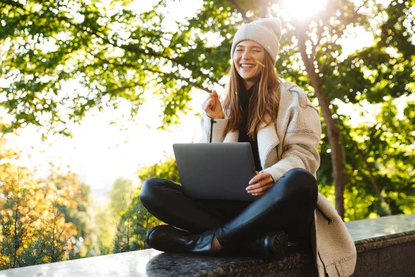 秋のコートと屋外に座って ラップトップ コンピューターを使用して 成功を祝っての帽子に身を包んだ美しい若い女性 — ストック写真