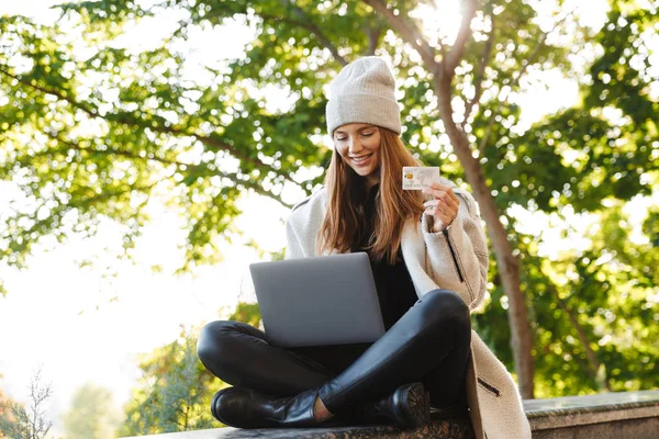 幸せな若い女を着た秋コートと帽子は屋外 ラップトップ コンピューターを使用して プラスチック製のクレジット カードを見せて — ストック写真