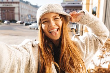 Bir selfie dilini yapışkanlık dışarı alarak sonbahar palto ve şapka açık, neşeli genç kadın giyinmiş