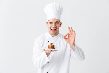 Cheerful man chef cook wearing uniform showing pastry on a plate isolated over white background