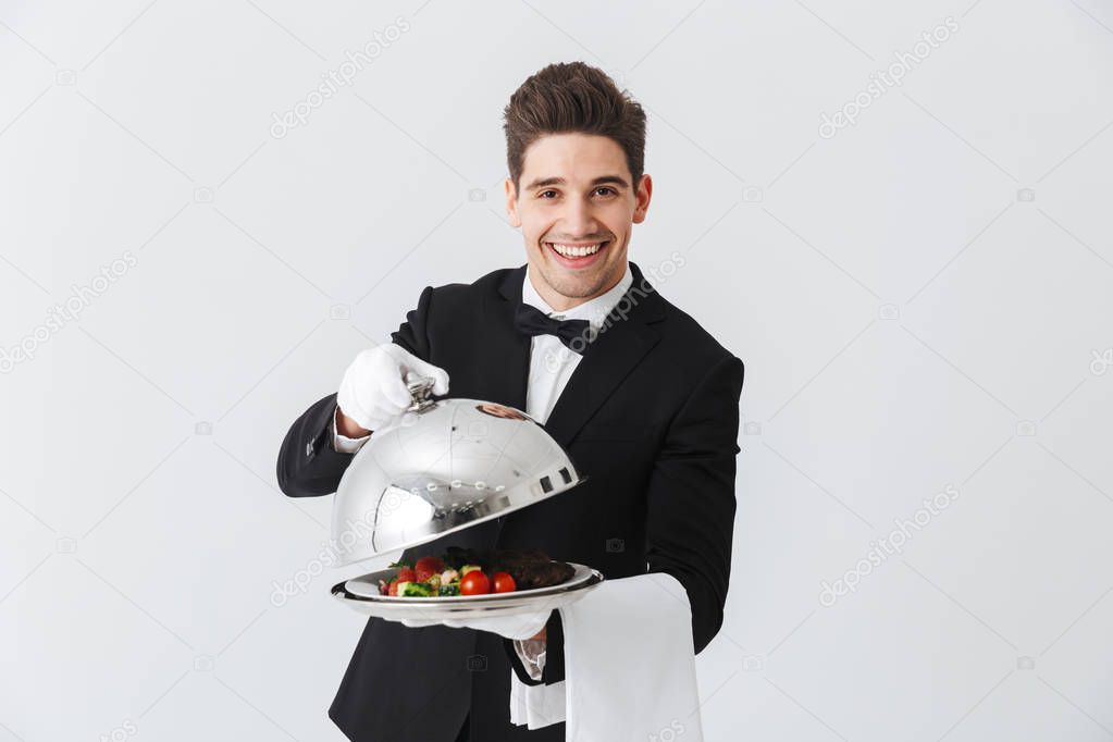 Portrait of a handsome young waiter in tuxedo showing beef steak dish on a plate over white background