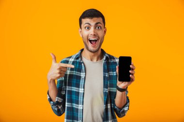 Boş ekran cep telefonu gösterilen turuncu arka plan üzerinde izole ekose gömlek ayakta giyen neşeli genç adam