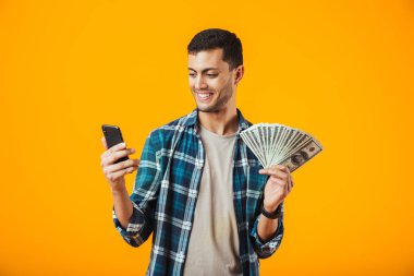 Cep telefonuyla para banknot gösterilen turuncu arka plan üzerinde izole ekose gömlek ayakta giyen neşeli genç adam