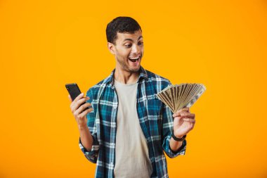 Cep telefonuyla para banknot gösterilen turuncu arka plan üzerinde izole ekose gömlek ayakta giyen neşeli genç adam